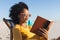 Happy african american woman in sunglasses sitting in deckchair reading book on sunny beach