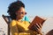 Happy african american woman in sunglasses sitting in deckchair reading book on sunny beach