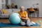 Happy aged woman resting on yoga mat with fitness ball, taking break from her domestic training, copy space