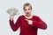 Happy amazed attractive young businesswoman in red blouse standing, holding many dollars, pointing finger, looking at camera with