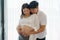 Happy Asian couple expecting baby standing together against window at home, loving husband tenderly touching belly of his pregnant