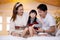 Happy asian family father and mother sitting teaching play ukulele guitar with daughter together on bed.