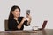 Happy Asian Korean businesswoman taking selfie photo with mobile phone at corporate company office desk smiling playful in female