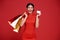 Happy Asian shopaholic woman wearing traditional cheongsam qipao dress holding credit card and shopping bag isolated on red