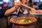 Happy Asian woman holding raw meat with chopsticks in shabu hotpot restaurant. Young girl making wow mouth eating dinner