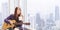 Happy Asian woman musician is holding guitar while sitting at the window with cup of coffee looking at the skyscraper and