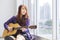 Happy Asian woman musician is holding guitar while sitting at the window looking out the skyscraper and cityscape view