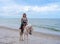 Happy Asian woman tourist in beachwear riding white horse on sand beach at Huahin beach with blue sea and cloudy sky