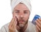 Happy and attractive camp homosexual man applying moisturizer facial cream with head wrapped in towel feeling confident