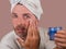 Happy and attractive camp homosexual man applying moisturizer facial cream with head wrapped in towel feeling confident