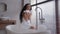 Happy attractive carefree hispanic woman sitting in bathroom naked in bath towel cheerful girl playing with soap blowing