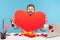 Happy bearded man looking behind big red paper heart and smiling, sitting at workplace with laptop in sticky hearts, showing his