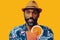 happy bearded mid adult african american man wearing Hawaiian shirt and hat smiling offering orange juice cocktail at