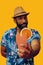 happy bearded mid adult african american man wearing Hawaiian shirt and hat smiling offering orange juice cocktail at
