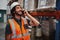 Happy bearded warehouse manager in conversation on mobile phone wearing white helmet and safety vest standing looking