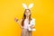 A happy beautiful girl on a head with rabbit ears holds Easter colored eggs and a plush rabbit on a yellow background