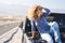 Happy beautiful mature woman in sunglasses posing with hand in curly hair while sitting on bonnet of jeep at highway. Cheerful