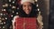 Happy beautiful mulatto girl in santas hat offering gift box, looking to camera and smiling standing at decorative