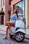 Happy beautiful woman with long brown hair dressed in trendy clothes posing with white retro Italian scooter on the