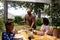 Happy biracial parents and daughter serving food and sitting at table for meal in garden