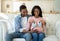 Happy black couple expecting new child, sitting on sofa together and holding baby booties