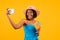 Happy black woman with yummy summer cocktail taking selfie on smartphone over orange studio background