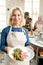 Happy blond mature woman in apron looking at you while going to serve table with selfmade food