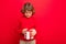 Happy blonde curly boy isolated over red background wall wearing red sweater holding gift box and looking at present box