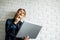 Happy blonde woman with laptop in her hands standing against white brick wall.