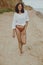 Happy boho girl in white shirt walking on sunny beach. Carefree stylish woman in swimsuit and shirt relaxing on seashore. Summer