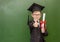 Happy boy with diploma in graduation cap stands near a school board and shows thumbs up gesture