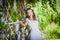 Happy bride in park posing near green fence. Wide angle shot of young woman in white wedding dress leans on hedgerow