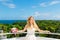 Happy bride standing next to the stone gazebo amid beautiful tropical landscape. Sea, sky, flowering plants and palm trees in