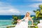 Happy bride standing next to the stone gazebo amid beautiful tropical landscape. Sea, sky, flowering plants and palm trees in