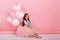 Happy brightful image of cute joyful little girl in tulle skirt sitting on present with balloons  on pink