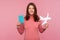 Happy brunette woman in pink sweater holding paper plane model and passport in hands looking at camera with toothy smile