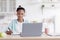 Happy busy teen black girl in earphones studying, doing homework at table with laptop in kitchen interior