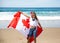 Happy Canadian girl carries fluttering white red flag of Canada against blue sky and ocean background.