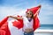 Happy Canadian girl carries fluttering white red flag of Canada against blue sky and ocean background.