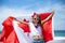 Happy Canadian girl carries fluttering white red flag of Canada against blue sky and ocean background.