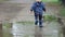 Happy carefree child runs through a puddle in raincoat and blue rubber boots