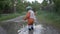 Happy carefree male baby in hat and rubber boots have fun playing with leaves in puddle on road after rain