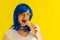 Happy Caucasian girl in a blue wig and sunglasses eating ice cream cone on a yellow background. Emotional excited woman