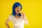 Happy Caucasian girl in a blue wig and sunglasses eating ice cream cone on a yellow background. Emotional excited woman