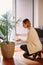 Happy Caucasian woman pouring water from glass jar on green home plant in pot. Large houseplant howea palm in apartment  house