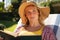 Happy caucasian woman wearing sunhat relaxing in sunny garden reading book, looking away, smiling