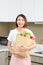 Happy, cheerful Asian woman holding paper bag full of fresh products, vegetables, greens, baguette and bananas