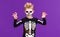 Happy cheerful boy in skeleton costume celebrates Halloween and scary gesture on purple background