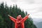 Happy and cheerful young male traveler wearing a red raincoat is raising his hands up, feeling great relief after climbing a