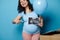 Happy cheery gravid woman smiling at camera, poiting at ultrasound image of her newborn baby, isolated blue background